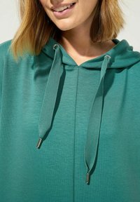 De teal hoodie heeft een gestructureerde stof met een ritssluiting aan de voorkant, een capuchon met dubbele trekkoorden en metalen accenten op de trekkoorden.