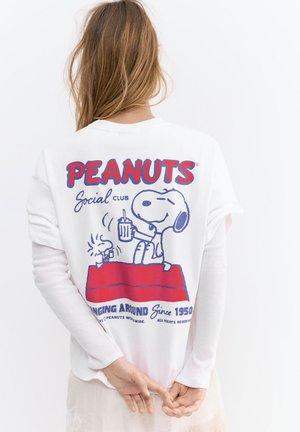 Osoba z długimi włosami nosząca białą koszulkę Peanuts z wizerunkiem Snoopy’ego i Woodstoka trzymających napoje na czerwonym psim budzie.