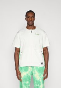 Λευκό μπουφάν με κοντά μανίκια, φερμουάρ μπροστά και δύο τσέπες με φερμουάρ· πράσινη διακόσμηση στα πλαϊνά. Συνδυάζεται με παντελόνι tie-dye σε πράσινη απόχρωση.