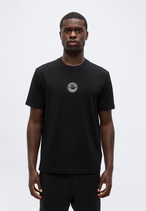 Noor mees musta T-särgiga, millel on väike ümar tenniseklubi logo rinna keskel, seisab lihtsa tausta ees.