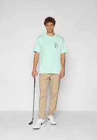 Myntegrøn t-shirt med et frontgrafik, parret med lysebeige bukser og hvide sneakers. Modellen holder en golfkølle.