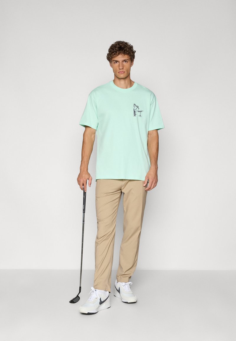 Mint green t-shirt with a front graphic, paired with light beige trousers and white sneakers. The model holds a golf club.