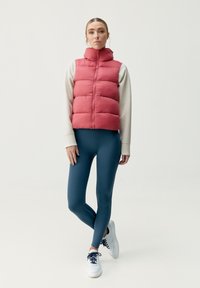 Röd puffervest med hög krage, dragkedja fram och horisontellt quiltat mönster, tillsammans med teal leggings och vita sneakers.