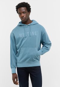 Hoodie bleu clair en coton doux, avec une poche kangourou à l'avant, une capuche avec cordon de serrage et un logo "MUSTANG" embossé sur la poitrine.