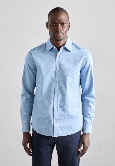 Light blue long-sleeve shirt made of cotton with a button-down collar, front button placket, and curved hem. Worn with dark trousers.