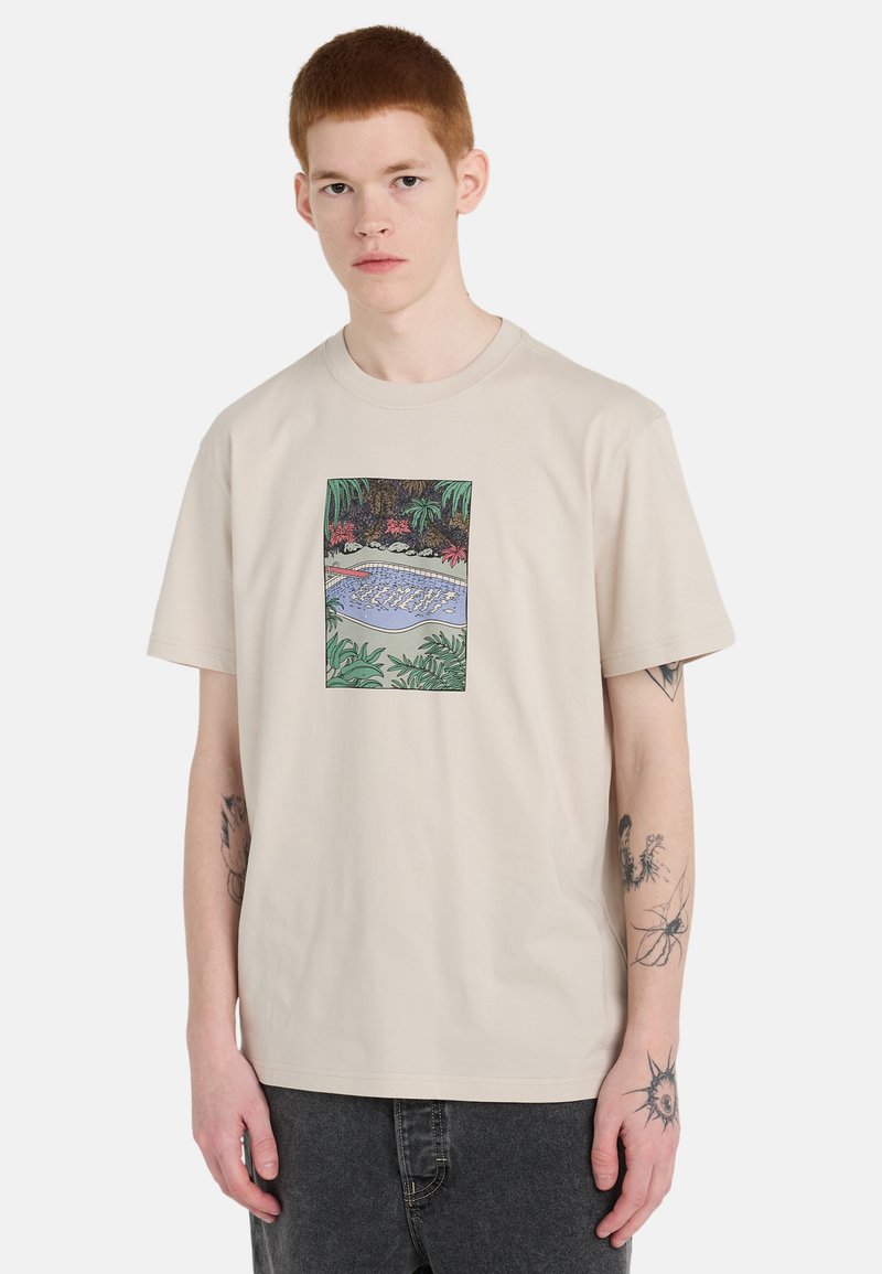 Junger Mann mit kurzen roten Haaren, der ein beiges T-Shirt mit einem bunten rechteckigen Pool- und Dschungelmotiv sowie dunkle Jeans trägt.