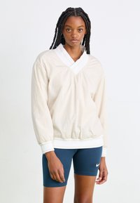 Μπεζ, ελαφρύ πουκάμισο με βαθύ V-λαιμό και λευκό γιακά. Διαθέτει ελαστικούς μανσέτες και στρίφωμα, με ένα διακριτικό λογότυπο στο στήθος.