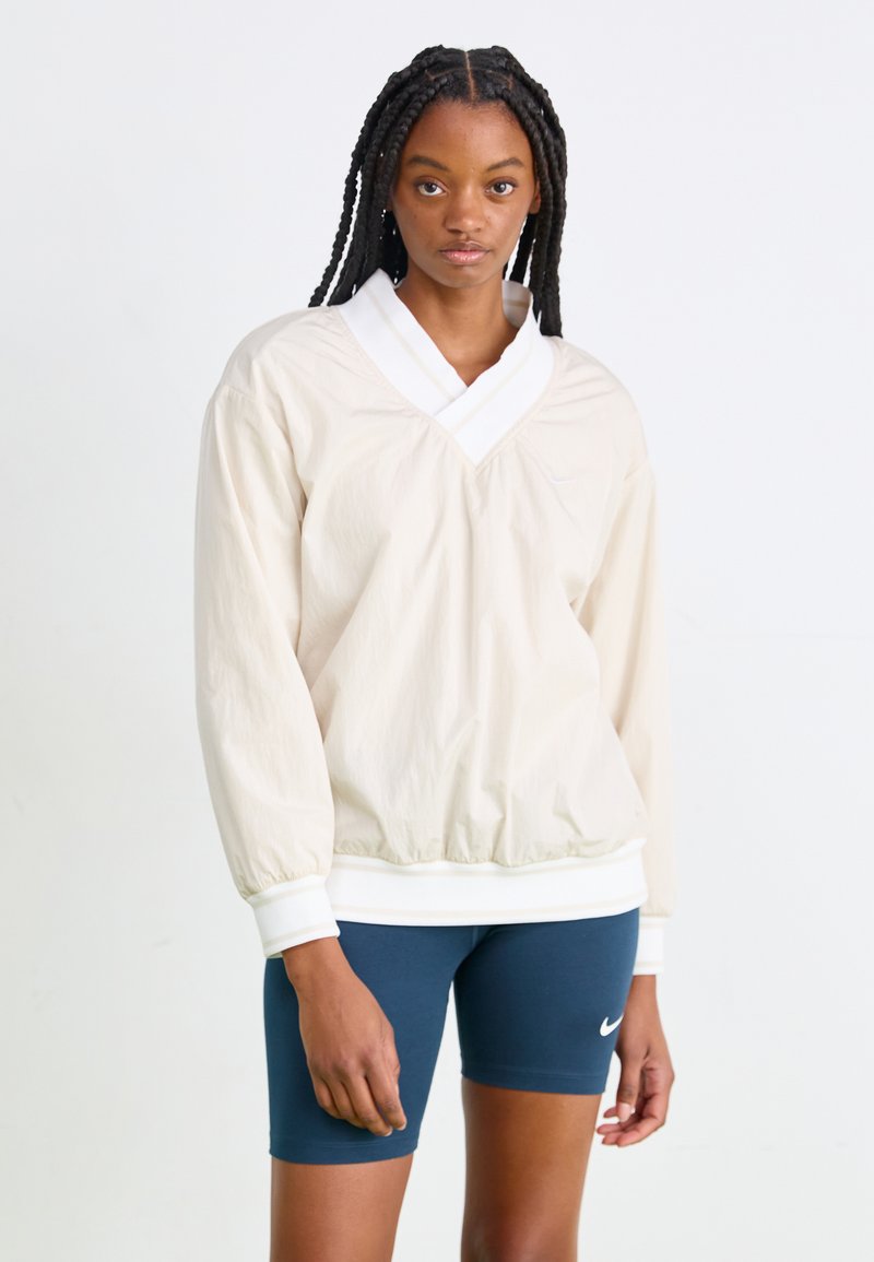 Μπεζ, ελαφρύ πουκάμισο με βαθύ V-λαιμό και λευκό γιακά. Διαθέτει ελαστικούς μανσέτες και στρίφωμα, με ένα διακριτικό λογότυπο στο στήθος.