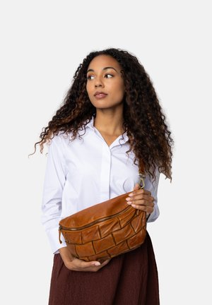 Sac en cuir brun à bandoulière avec un design matelassé et une fermeture éclair, tenu contre une chemise blanche. Matériau durable avec une texture douce.