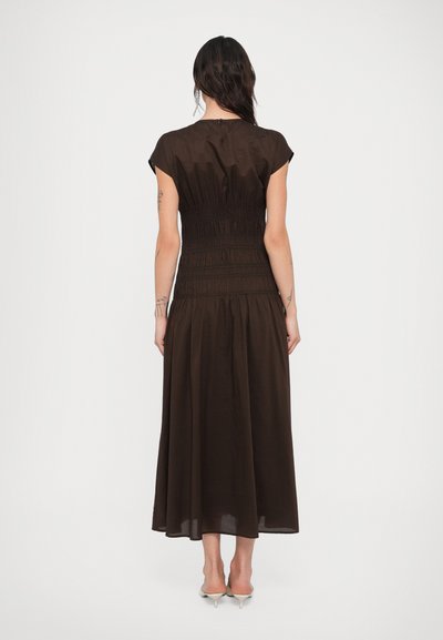 Γυναίκα που φαίνεται από πίσω, φοράει ένα σκούρο καφέ φόρεμα μέσης μήκους με κοντά μανίκια και σφηκοειδή ζώνη στη μέση, καθώς και μπεζ παπούτσια με χαμηλό τακούνι, μπροστά σε λευκό φόντο.