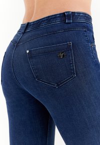 Dark blue denim jeans with a fitted design, featuring a back pocket with a small black logo and subtle stitching details.