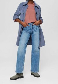 Langer, hellblauer Mantel über einer rosafarbenen Bluse, kombiniert mit hellblauen, weit geschnittenen Jeans und grauen Stiefeletten.