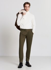 Weißer Rollkragenpullover aus Strick, olivgrüne Hosen mit zwei Fronttaschen und schwarze Stiefeletten. Model hält eine braune Jacke in der Hand.