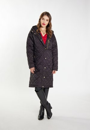 Zwarte gevoerde lange jas met capuchon, voorzien van gouden drukknoopjes, een diagonale ritszak en een cinched taille. G gedragen over een rode top en zwarte laarzen.