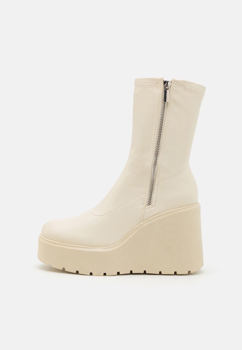 Botte compensée mi-mollet couleur crème avec semelle plateforme épaisse et fermeture éclair latérale, texture lisse, et bout arrondi.