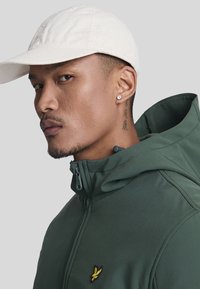 Groene capuchonjack met een ritssluiting, voorzien van een klein geel geborduurd logo. Gekocht met een witte pet en een stud oorbel. Gladde textuur.