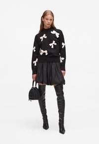 Czarny sweter z białymi wzorami w kokardy, zestawiony z czarną rozkloszowaną spódnicą. Modelka trzyma czarną plecioną torbę, nosi czarne wysokie kozaki.
