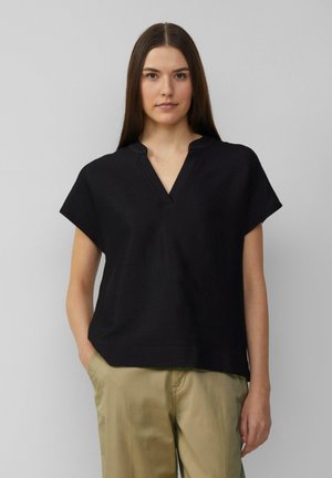 Zwarte top met korte mouwen en een gestructureerd ribbeloppervlak, V-hals en een ontspannen pasvorm. In combinatie met lichtgekleurde broeken.