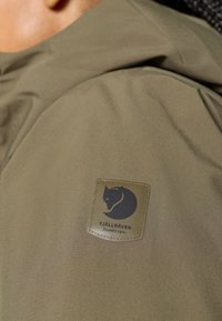 Maslinasto zeleni jakna s glatkom teksturom koja sadrži kožnu zakrpu s logotipom crnog zeca i tekstom "FJÄLLRÄVEN Sweden 1960."