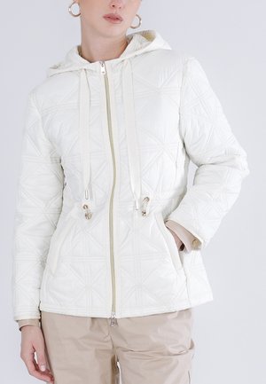Kvinde iført en hvid quiltet jakke med lynlås og hætte samt beige bukser, med hånden i lommen, foran en ensartet lys baggrund.