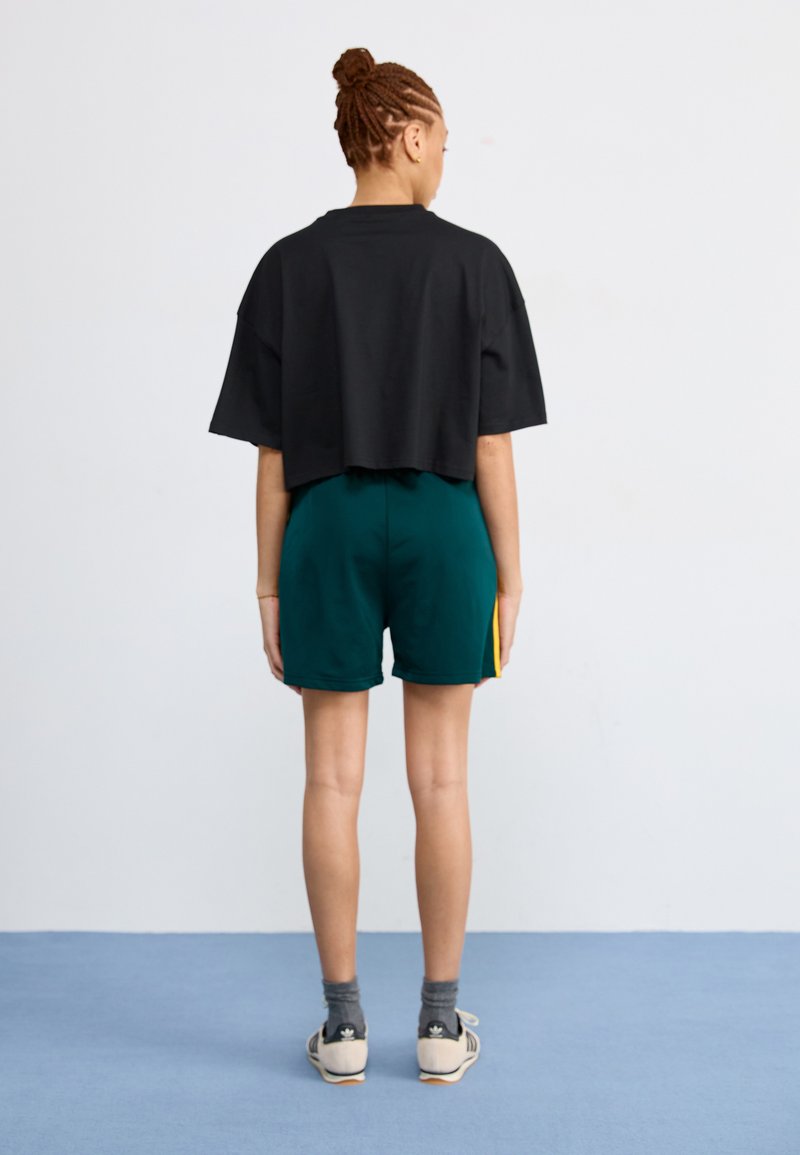 Svart kortärmad tröja, gröna shorts med gul sidosträck, grå strumpor och vita skor. Tagen bakifrån mot en ljus bakgrund.