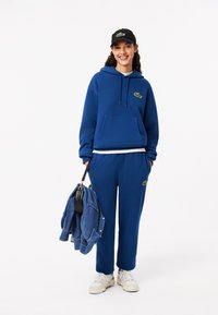 Blauwe hoodie en broek set met groene logo-details, gemaakt van een katoenmix, casual fitting, gecombineerd met een zwarte pet en spijkerjack.