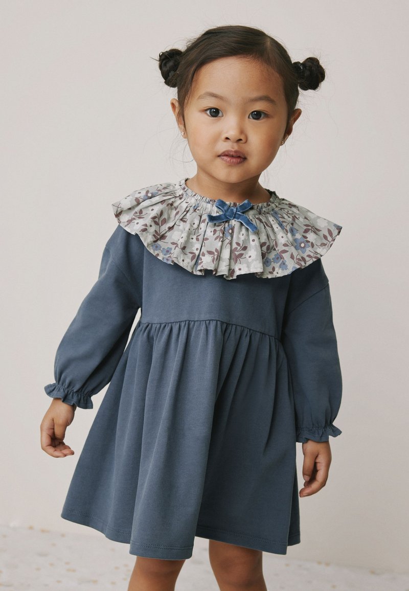 Modré šaty s dlouhými rukávy a nařaseným pasem, s květovaným volánkovým límcem a mašlí u výstřihu. Měkký bavlněný materiál.