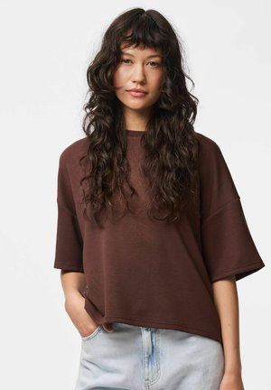 Ung kvinde med langt, krøllet mørkt hår iført en løs brun kortærmet top og lyseblå højtaljede jeans, hånd i lommen.