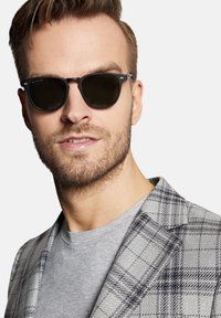 Okrągłe okulary przeciwsłoneczne z ciemnymi soczewkami, czarną oprawką, srebrnymi akcentami, w zestawie z szarym t-shirtem i szarym wierzchnim ubraniem w kratę. Widoczna krótka broda.