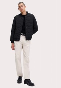 Svart quiltad jacka med vågmönster, bärs över en svart skjorta. Kombinerad med gräddfärgade byxor och svarta snörskor.