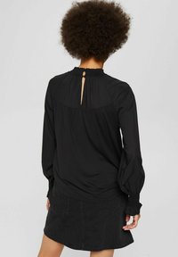 Sort langærmet bluse med en indsamlet halsudskæring og detaljer med nøglehul bagpå, udstyret med elastiske manchetter og blød tekstur. Parret med en sort denimnederdel.
