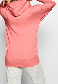 Sweat à capuche corail à texture lisse, avec une poche kangourou et un ourlet côtelé, associé à un pantalon de survêtement gris clair à motifs.