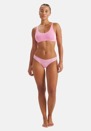 Nainen seisoo paljain jaloin, yllään vaaleanpunainen urheilurintaliivit ja yhteen sopivat bikini-tyyliset alushousut, yksinkertaista valkoista taustaa vasten.