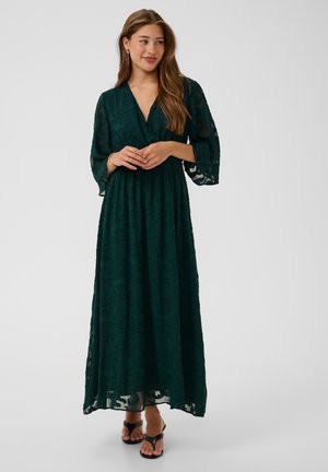 Femme portant une longue robe vert foncé à motifs avec des manches transparentes et des talons ouverts noirs, debout et regardant sur le côté.