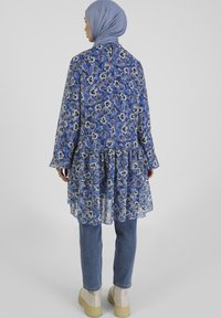 Niebieska kwiatowa długa bluzka z przezroczystego materiału, z falbaniastym dołem i długimi rękawami, w zestawieniu z jasnoniebieskimi dżinsami i beżowymi botkami za kostkę.