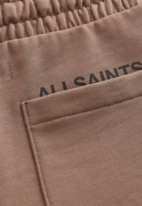 Detailní záběr hnědého látkového pásku a kapsy s černým částečným textem "ALL SAINTS" vytištěným na měkkém texturovaném materiálu.