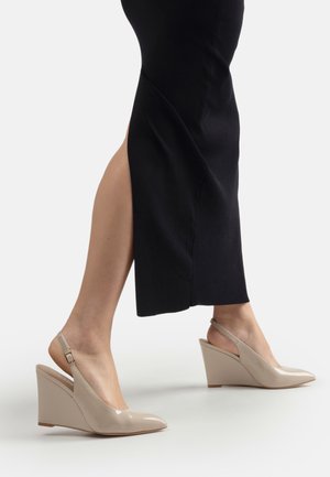 Décolleté beige in vernice con zeppa e cinturino posteriore, indossate con un abito nero con uno spacco laterale alto, viste dalla metà del polpaccio in giù.