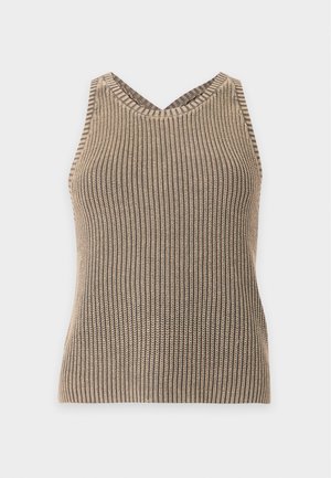 Vrouw met zwarte zonnebril, beige gebreide tanktop, wit ondershirt, brede vervaagde groene broek en zwarte puntige schoenen, staand met handen in de zakken.