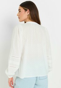 Blusa blanca de manga larga con lunares texturizados, detalles de pliegues en las mangas y un ajuste holgado. Presenta un escote redondo y puños elásticos.