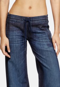 Pantalons joggers en denim bleu foncé avec taille élastique, jambes larges et délavage subtil. Présente un patch logo sur le devant à gauche.