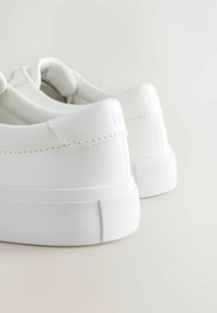 Tenisi albi din material sintetic neted, cu un design joasă la gleznă, talpă din cauciuc și detalii subtile de cusătură la călcâi și pe laturi.