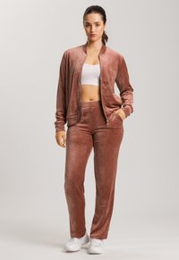 Sametový sportovní set v jemně růžové barvě, skládající se z bomber bundy na zip a odpovídajících kalhot s elastickým pasem. Look doplňují bílé sportovní boty.