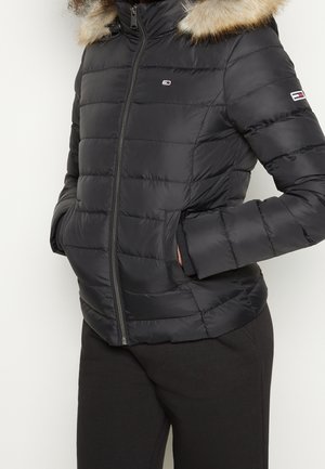 Veste matelassée noire avec capuche bordée de fourrure, fermeture éclair à l'avant, poches latérales et petits logos de la marque sur la poitrine et la manche, portée avec un pantalon noir.
