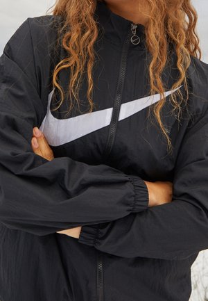 Personne aux longs cheveux bouclés portant une veste zippée noire Nike avec un grand logo swoosh blanc, les bras croisés sur la poitrine.
