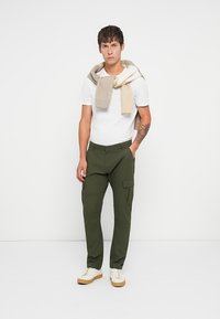 Olivgrüne Cargo-Hosen mit Seitentaschen, kombiniert mit einem weißen T-Shirt und einem beigefarbenen Pullover, der über die Schultern drapiert ist. Legere Sneakers.