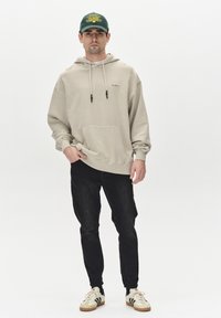 Beige hættetrøje med frontlomme, snore og logo, parret med sorte jeans og beige sneakers med sorte striber og snørebånd.