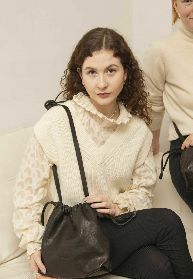 Maglione lavorato a maglia color crema con scollo a V sopra una blusa trasparente con volant. Borsa a tracolla in pelle nera con finitura lucida e texture liscia.