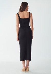 Black fitted dress with a square neckline, thick straps, and a back zipper. Midi length with a smooth texture, worn with open-toed heels.