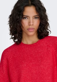 Pull rouge en maille avec un col ras du cou, des poignets côtelés et une coupe décontractée. Tissu texturé avec une légère variation de couleur.