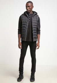 Vesta neagră căptușită cu glugă, purtată peste un tricou cu mânecă lungă henley negru, asortată cu blugi skinny negri și adidași negri.